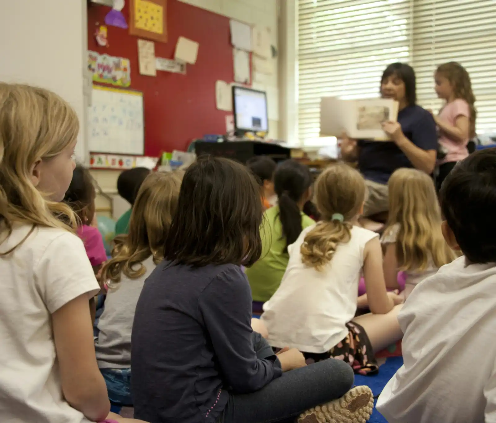 Children sitting on the floor in a classroom as a teacher reads a story to them.